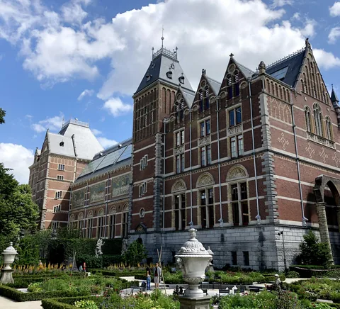 Real Rijksmuseum back/side garden exterior. Natural colours, architectural/garden visitor context, no removed I amsterdam sign or dominant temporary exhibition poster. Near-square crop focuses right historical tower a…