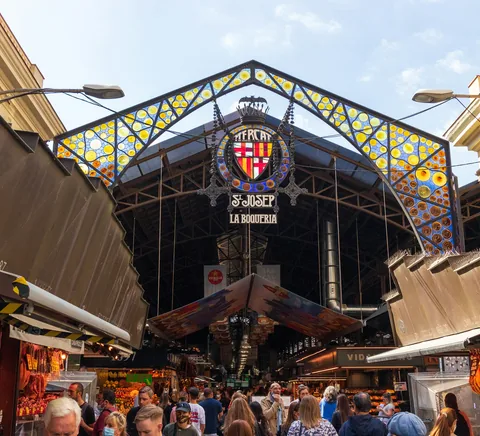Busy La Boqueria entrance with food stalls, market shoppers and coloured canopy.