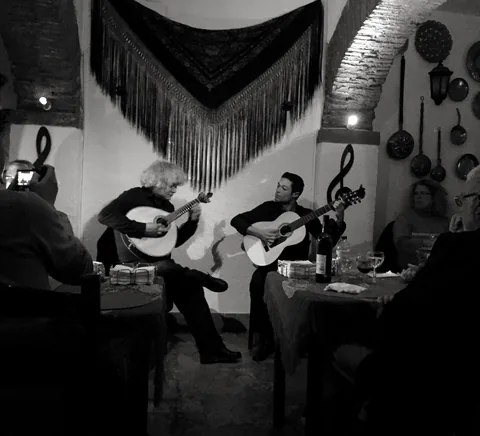Two musicians with Portuguese guitar and guitar in an intimate Alfama fado room, audience close by; monochrome documentary photograph.